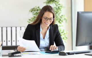 Smiling businesswoman wearing glasses, reviewing documents and using a calculator at her desk in a modern office.