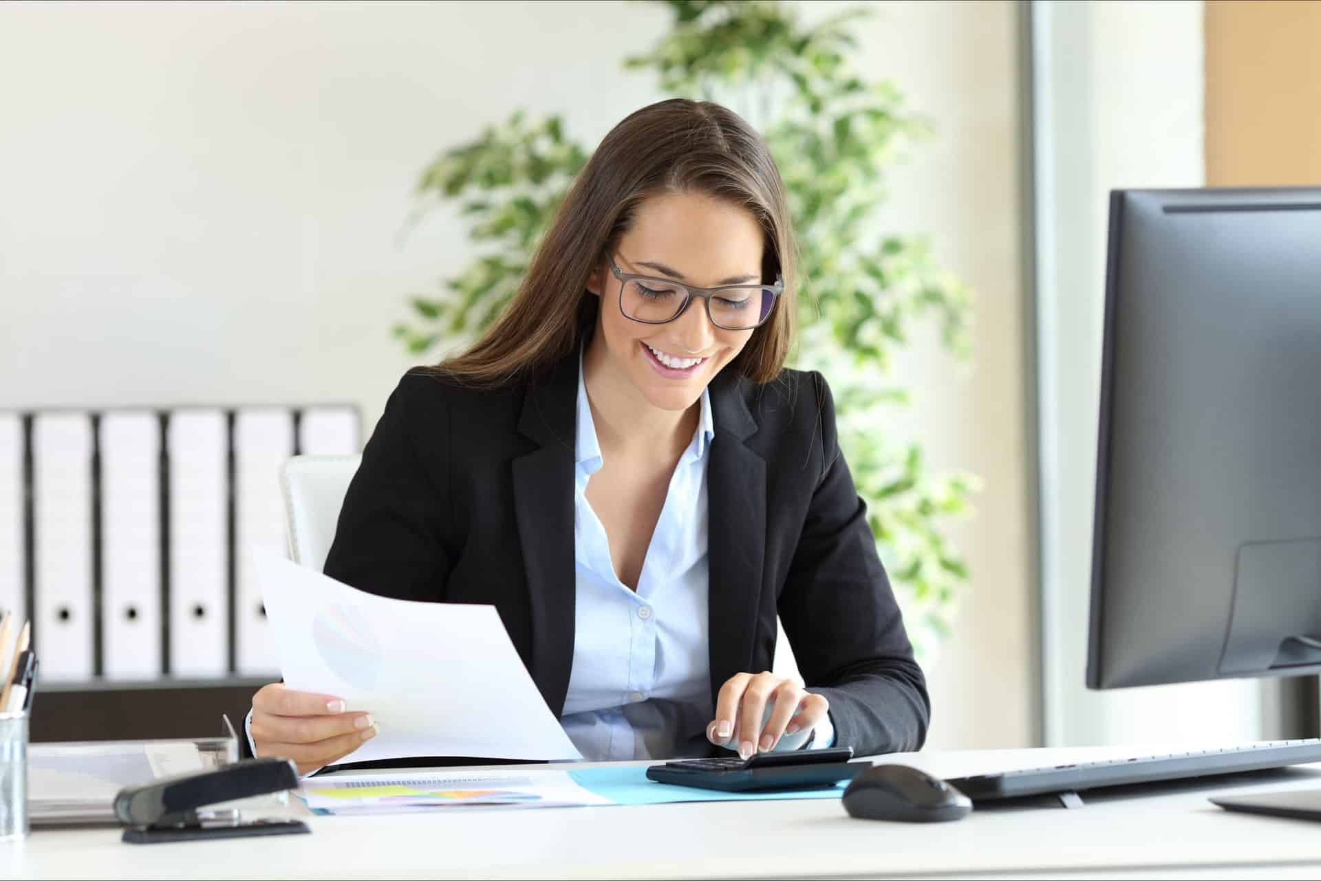 Smiling businesswoman wearing glasses, reviewing documents and using a calculator at her desk in a modern office.